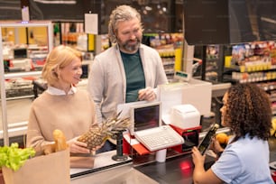 Pareja madura parada junto al mostrador del cajero en el supermercado mientras una joven de raza mixta escanea los productos alimenticios que compraron