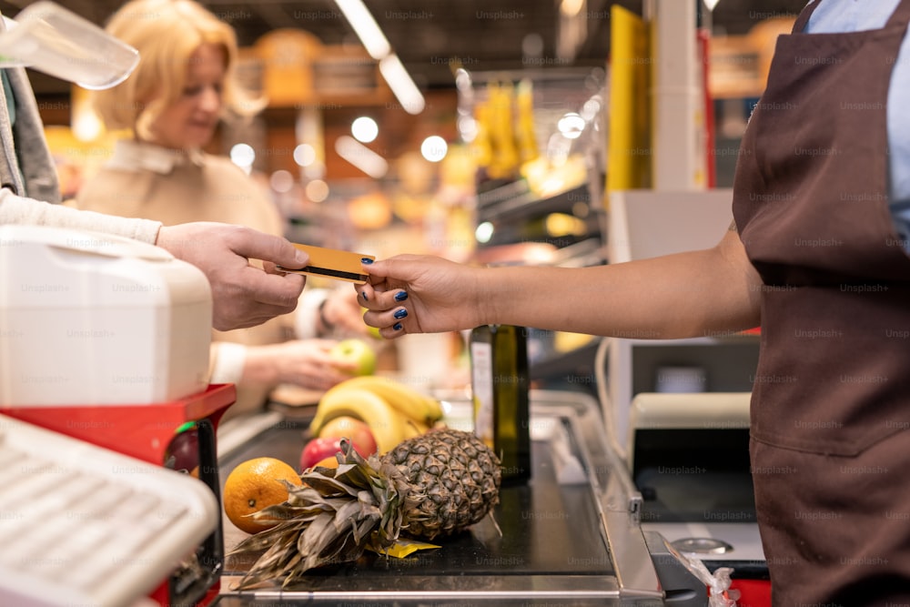 갈색 앞치마를 입은 현대 계산원은 슈퍼마켓에서 서빙하는 동안 현금 상자를 통해 성숙한 고객의 신용 카드를 돌려주거나 가져갑니다