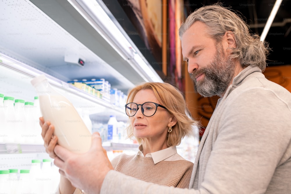 Aged serious bearded man and his blond wife choosing milk on display with dairy products while standing by shelves with bottles