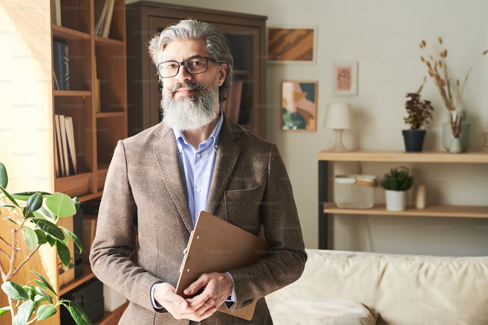 Elegant bearded aged professional in eyeglasses and formalwear standing in front of camera on background of shelves and couch