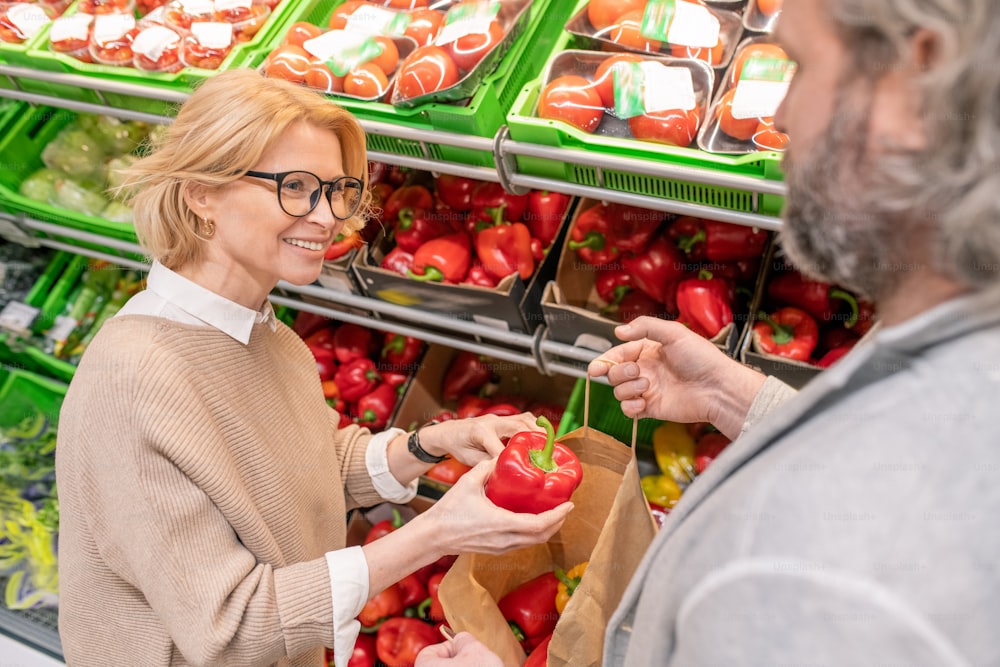 Happy blond woman choosing red ripe capsicum on display with fresh vegetables and putting it into paperbag held by her husband