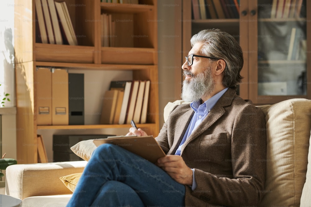 Profissional de idade séria com cabelos grisalhos e barba sentada no sofá em frente à janela e fazendo anotações no fundo de prateleiras com livros
