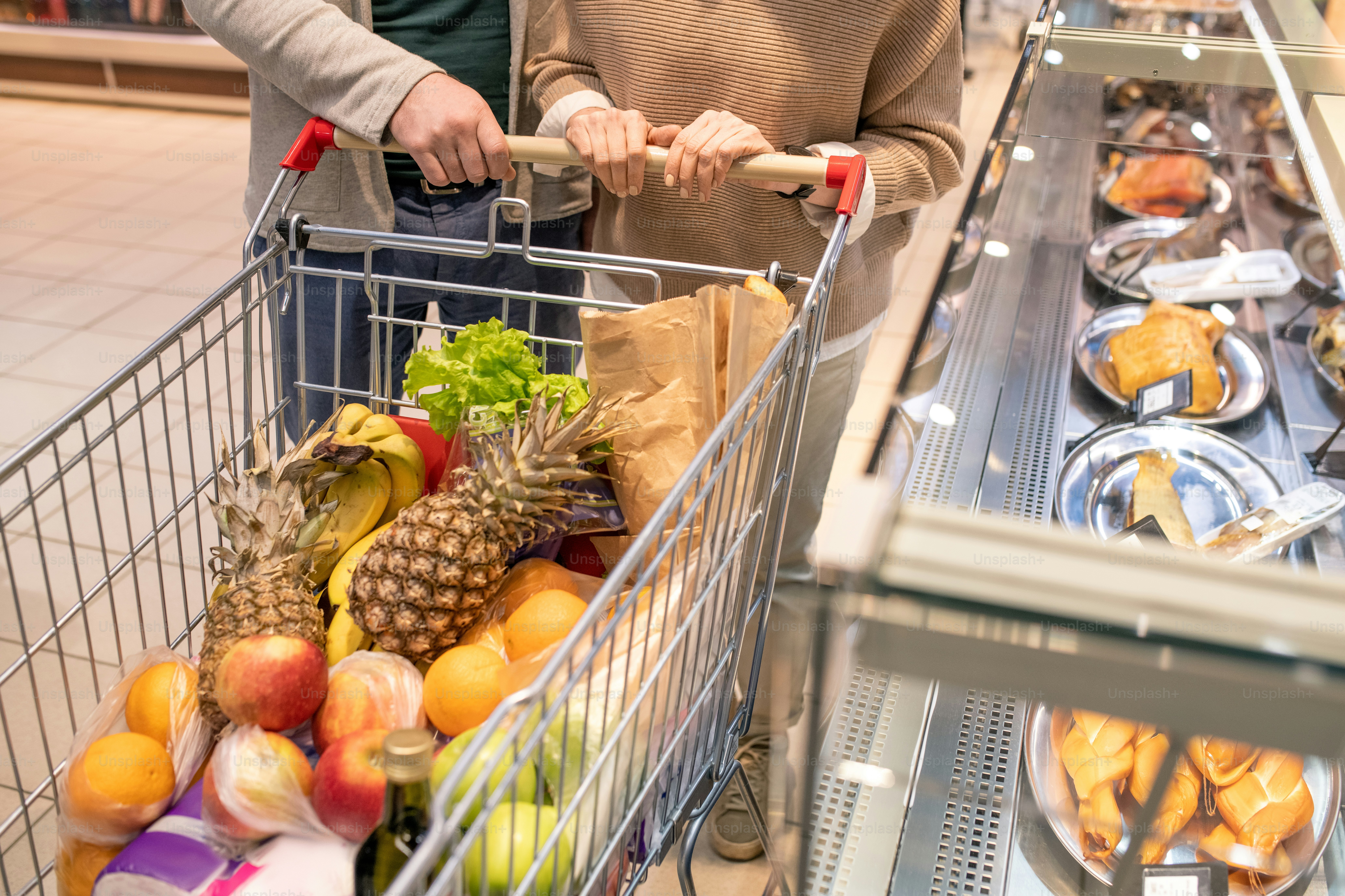 Hands of contemporary mature couple pushing shopping cart with fresh food products while passing by large display with grilled chicken