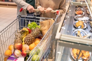 Hands of contemporary mature couple pushing shopping cart with fresh food products while passing by large display with grilled chicken