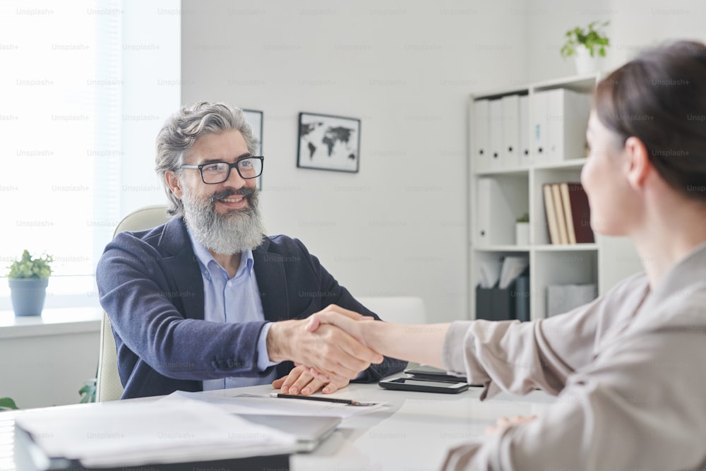 Smiling mature employer with grey beard and hair shaking hand of young female over desk after interview and signing working contract