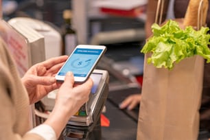 Manos de una clienta madura contemporánea con teléfono inteligente sobre máquina de pago de pie junto al mostrador del cajero en el supermercado