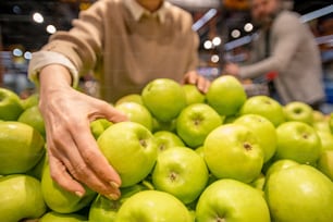 Hand einer reifen Verbraucherin, die frischen Granny-Smith-Apfel nimmt, während sie während des Besuchs im Supermarkt neben einem Haufen Obst steht