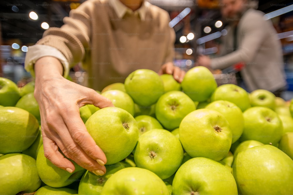 Mano de consumidora madura tomando manzana fresca granny smith mientras está de pie junto a un montón de fruta durante la visita al supermercado