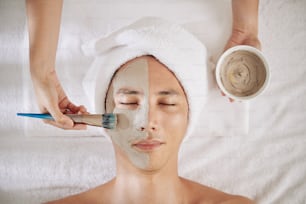 Young man getting anti-aging clay mask applied on his face in beauty salon