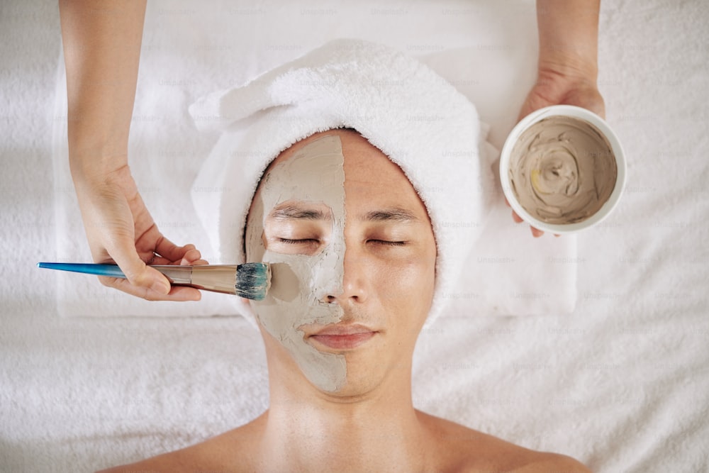 Junger Mann, der im Schönheitssalon eine Anti-Aging-Tonmaske auf sein Gesicht aufgetragen bekommt