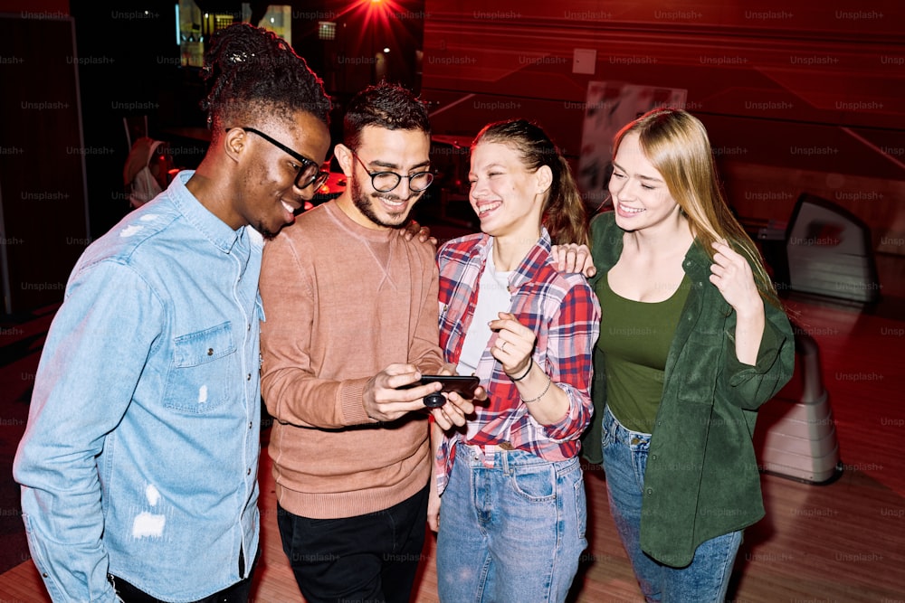 Young smiling man with smartphone showing friends photo of his girlfriend taken by him during game of bowling in leisure center