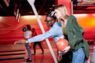 Jeune homme d’origine africaine pointant du doigt la piste de bowling tout en expliquant à sa petite amie comment lancer une balle pour frapper toutes les quilles