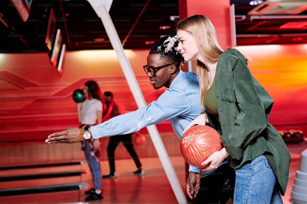 Junger Mann afrikanischer Abstammung, der auf die Bowlingbahn zeigt, während er seiner Freundin erklärt, wie man einen Ball wirft, um alle Kegel zu schlagen