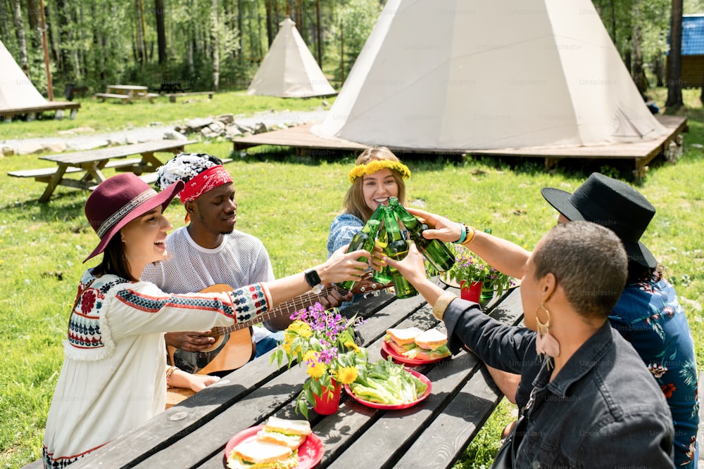 Group of positive multi-ethnic friends sitting at table with snacks and drinking beer together at campsite