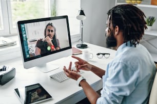 Joven africano ocupado en camisa hablando con su compañero de trabajo usando la computadora mientras está sentado en el interior