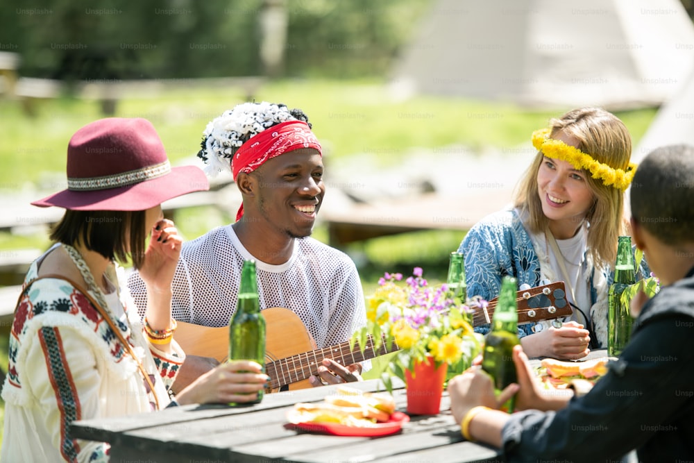 Smiling Afro-American musician playing guitar while sitting with friends at one table outdoors