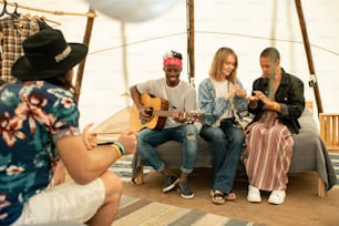 Grupo de jóvenes amigos multiétnicos sentados en una tienda de campaña y escuchando música de guitarra interpretada por un chico negro