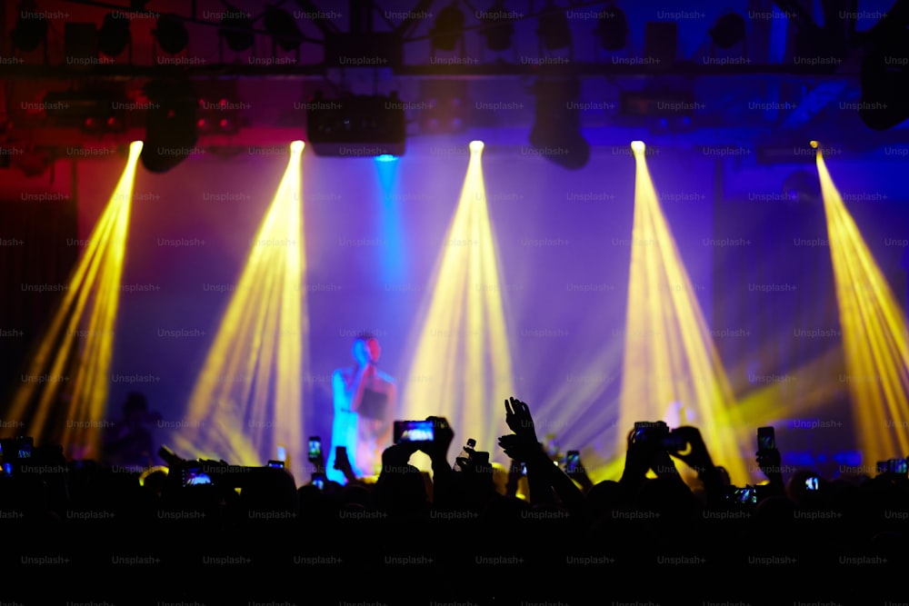 Fans in crowd raising hands up cheering in front of stage at music concert, admiring great performance of young singer in bright lights and filming it with smartphones