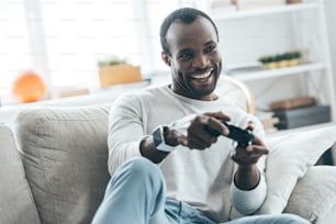 Bel giovane africano che gioca ai videogiochi e sorride mentre è seduto sul divano di casa