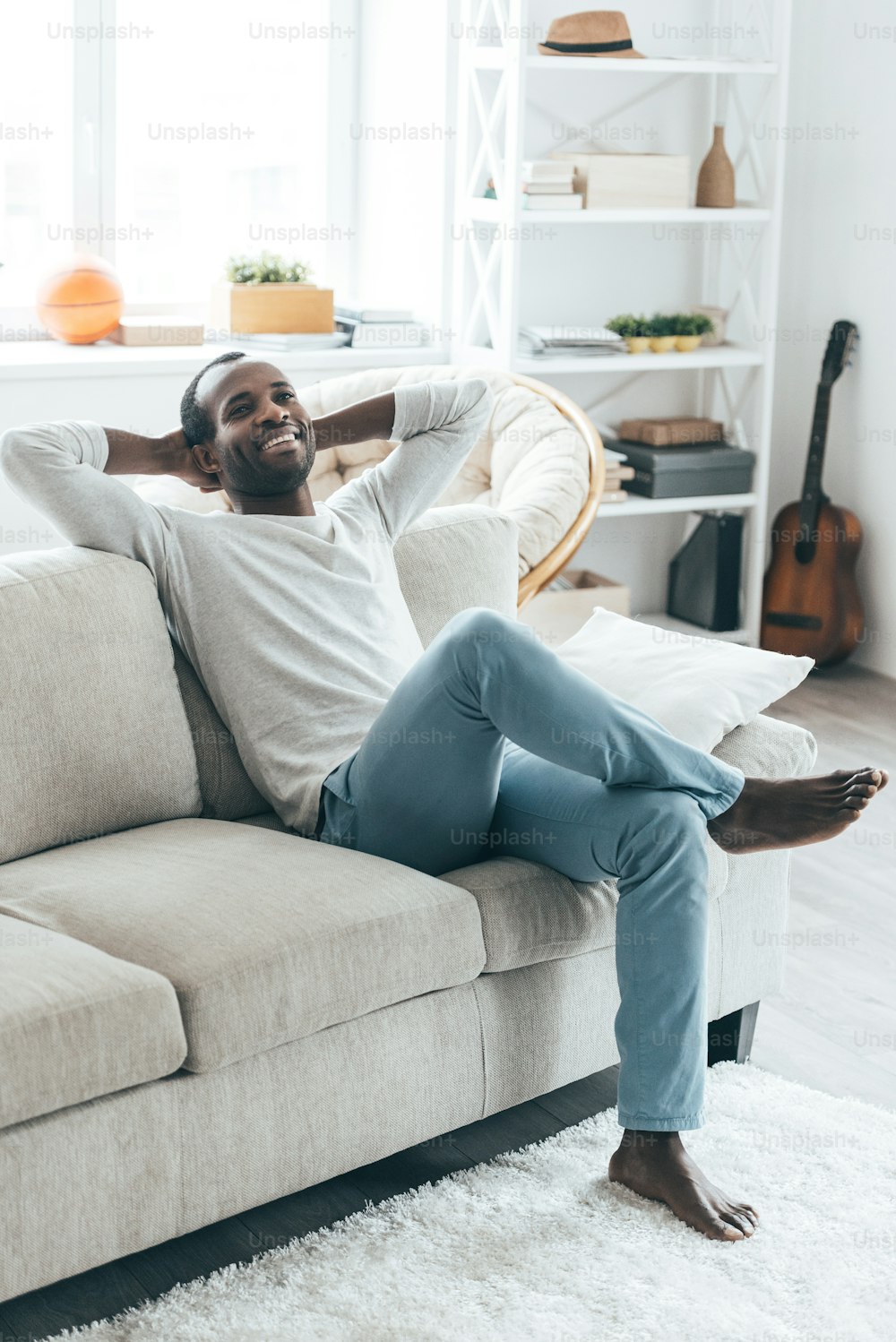 Apuesto joven africano con las manos detrás de la cabeza y sonriendo mientras está sentado en el sofá de su casa