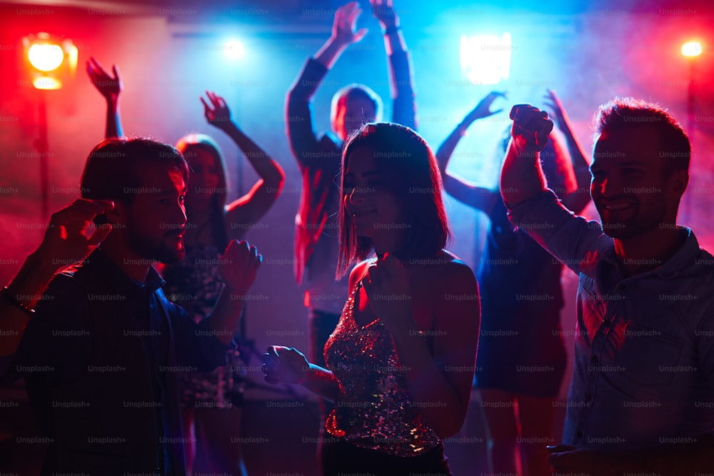 Giovane ragazza graziosa in top splendente ballando con amici maschi alla festa in discoteca studentesca e godendo della musica con gli occhi chiusi