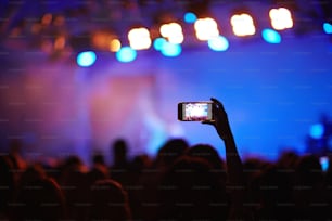 Back lit image of crowd at music concert before stage, focus on one fan holding cellphone high above heads to take picture of memorable moment