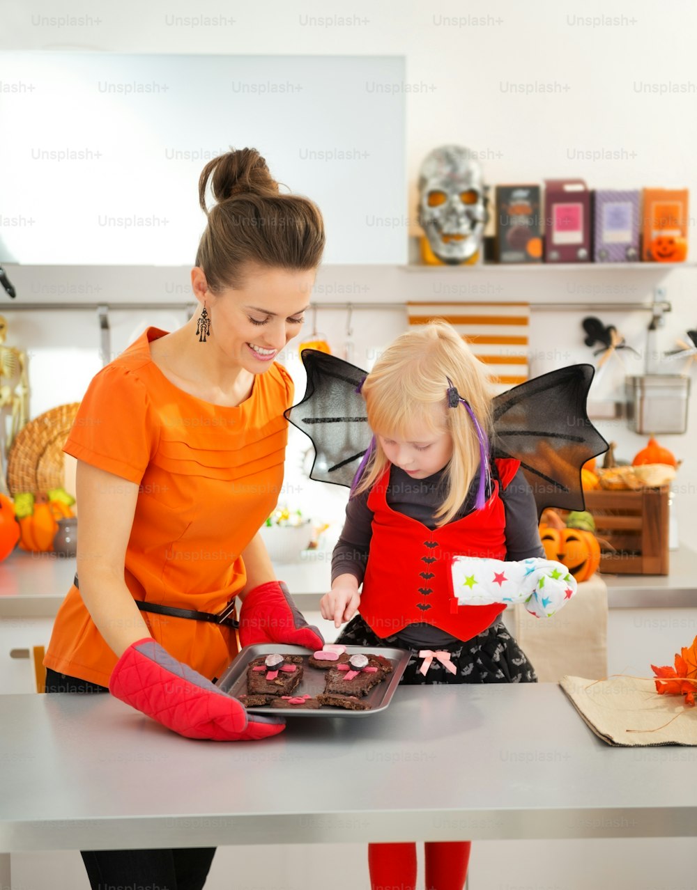 Halloween dressed girl with smiling young mother in decorated kitchen holding tray with freshly baked Halloween biscuits for Trick or Treat. Ready to halloween party. Traditional autumn holiday