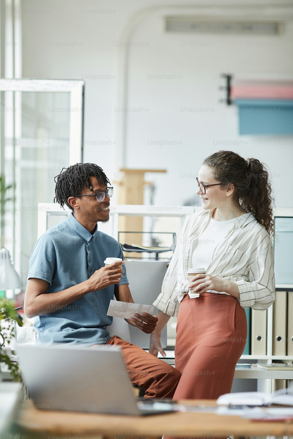 Vertical portrait of two creative young people looking at printed photographs and chatting during coffee break in modern office