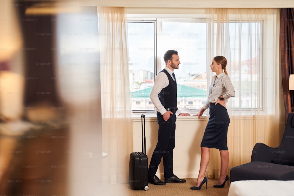 Young elegant male and female are standing at window in hotel room with luggage and communicating