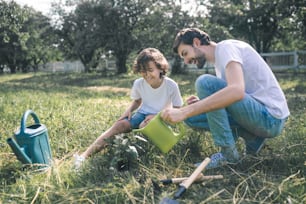 Gardening. Dark-haired man holding the watering can and watering the plants in the garden, his son watching him