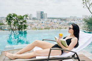 Mulher jovem muito magra relaxando no chaise-lounge perto da piscina infinita com livro e copo de juce laranja e olhando para a cidade grande