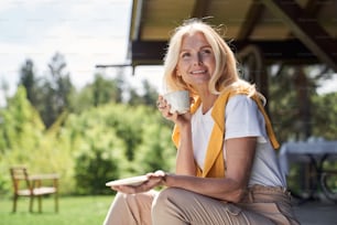 Bajo ángulo de sonriente atractiva mujer sentada en la terraza de la casa de campo y tomando una taza de café