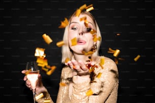 Jeune belle femme aux cheveux blonds avec une flûte de champagne soufflant des confettis dorés de sa main tout en profitant de la fête