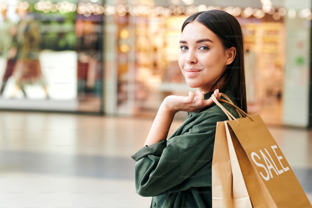 Hübscher junger brünetter Shopaholic mit Papiertüte auf der Schulter, der während der Verkaufssaison vor der Kamera in einem großen modernen Handelszentrum steht