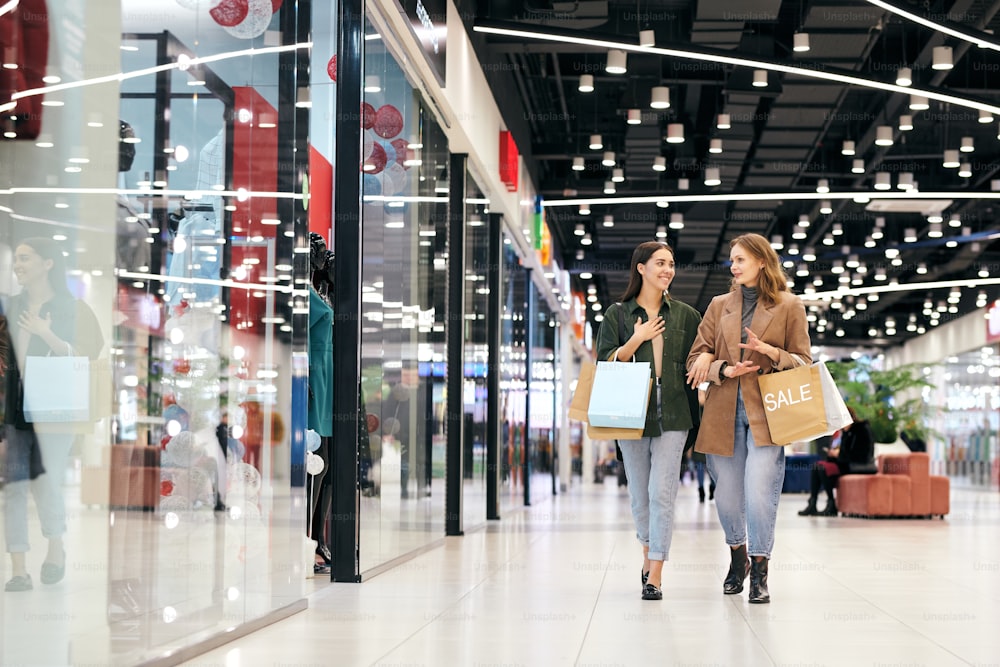 우아한 캐주얼웨어를 입은 두 명의 젊은 현대 여성이 쇼핑몰 안의 상점 창문을 따라 걷고 무엇을 사고 싶은지 논의합니다