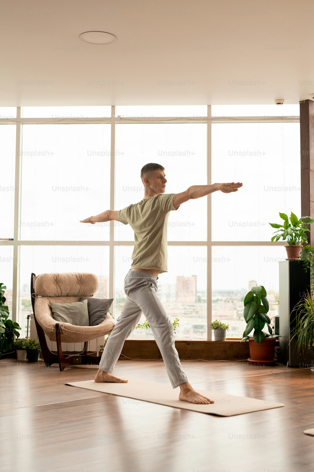 Joven activo en ropa deportiva de pie sobre una colchoneta con los brazos extendidos durante el entrenamiento de yoga en el entorno del hogar