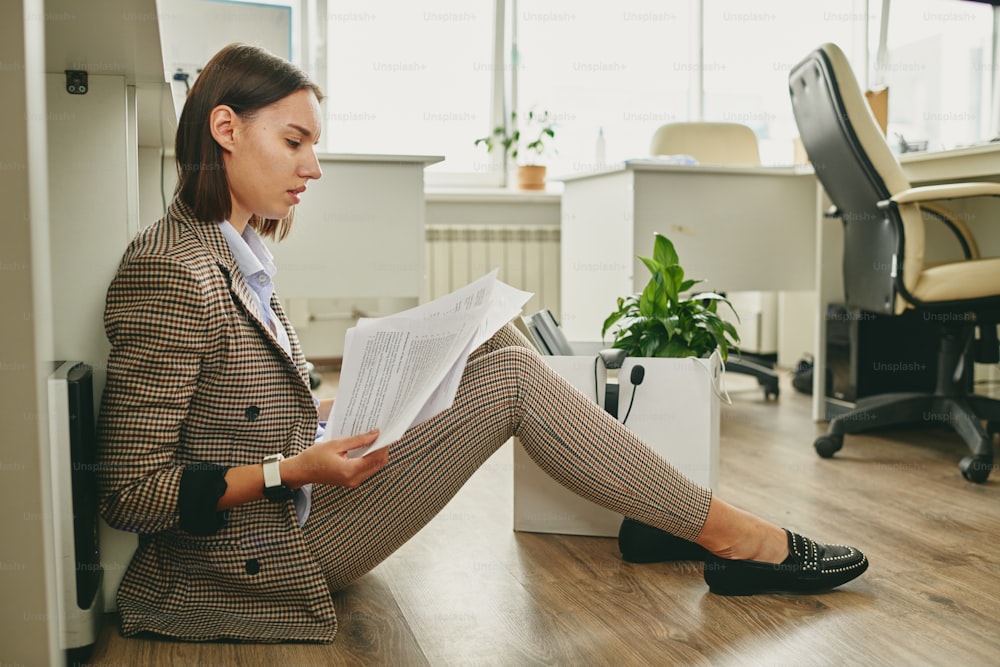 Young contemporary businesswoman in smart casualwear sitting on the floor by desk and looking through financial papers in office