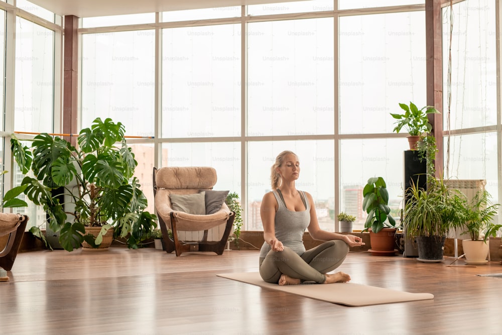 スポーツウェアを着た若い穏やかな金髪の女性が足を組んで、蓮のポーズでマットの上に座り、家で瞑想の練習をする