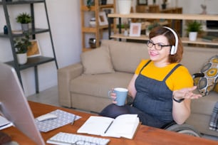 Contemporary young disable female student in headphones having drink and talking with teacher during online lesson in front of computer