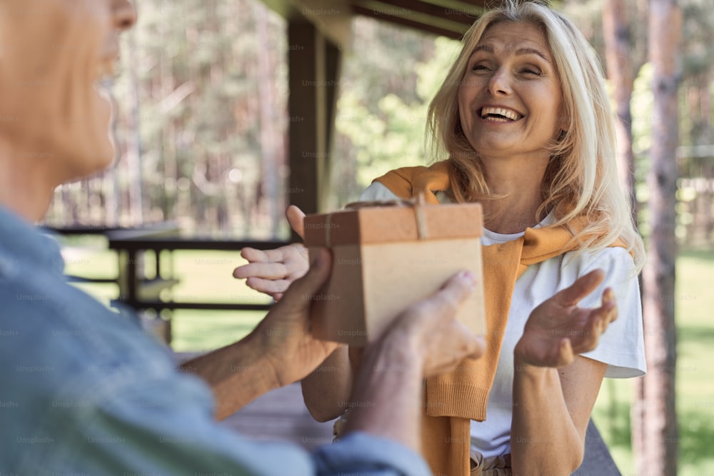 Mulher alegre está se sentindo surpresa enquanto homem está dando sua caixa com presente na varanda ao ar livre