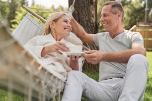 Mulher feliz com xícara de chá deitada na rede e conversando com o marido ao ar livre