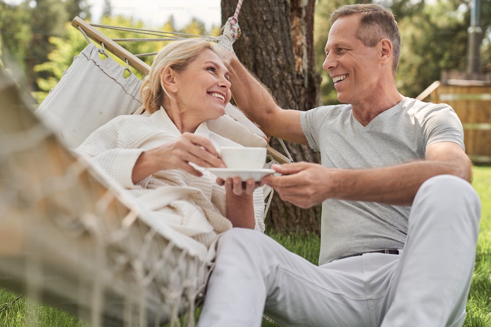 Happy woman with cup of tea lying in hammock and talking to husband in open air