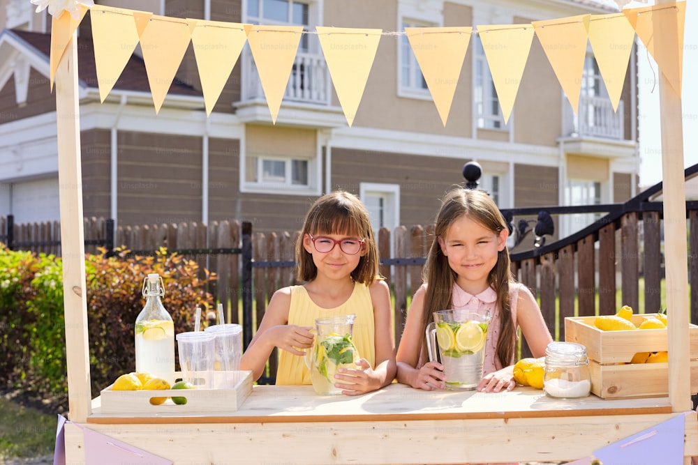 Dos niñas felices y adorables de pie junto a un puesto de madera decorado con pequeñas banderas y vendiendo limonada casera fresca al aire libre