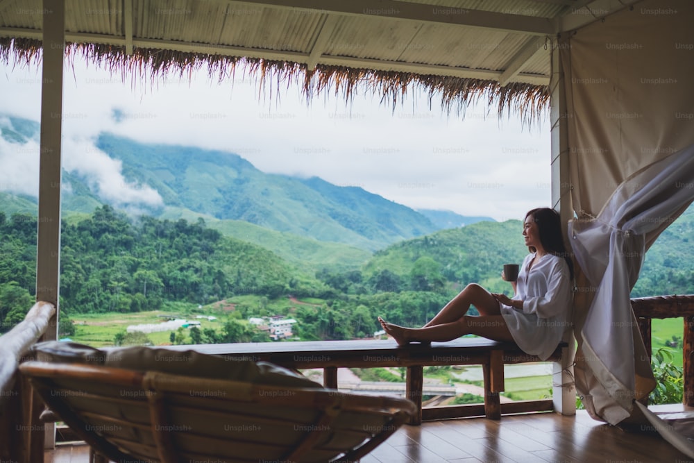 Portrait image of a beautiful asian woman holding and drinking hot coffee on balcony , looking at mountains and green nature
