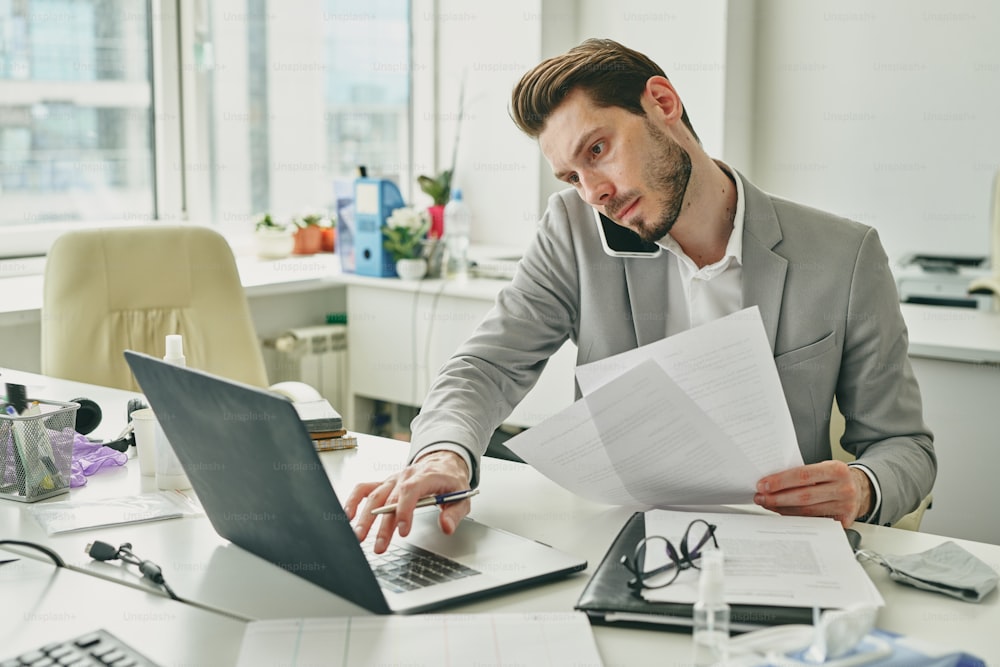 Busy young office worker in formalwear consulting client on mobile phone while looking at data on laptop display and reading financial papers