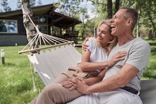 Merry mature man is hugging wife as they are sitting in hammock together and enjoying sunny day outdoors