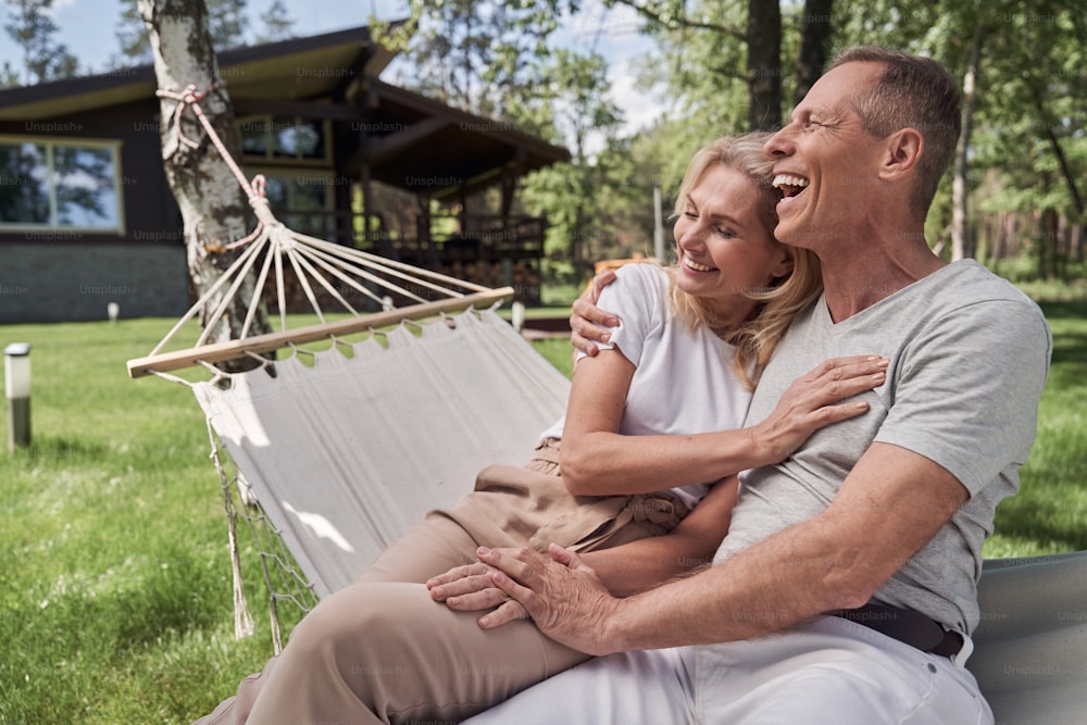 Merry mature man is hugging wife as they are sitting in hammock together and enjoying sunny day outdoors