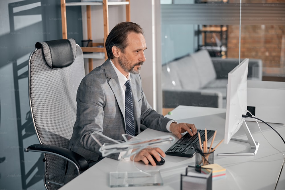 Bearded gentleman in gray suit working on desktop PC while sitting at the table in office