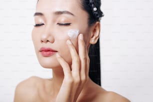 Giovane donna asiatica che chiude gli occhi e applica la crema morbida mosturizzante anti-invecchiamento sul suo viso