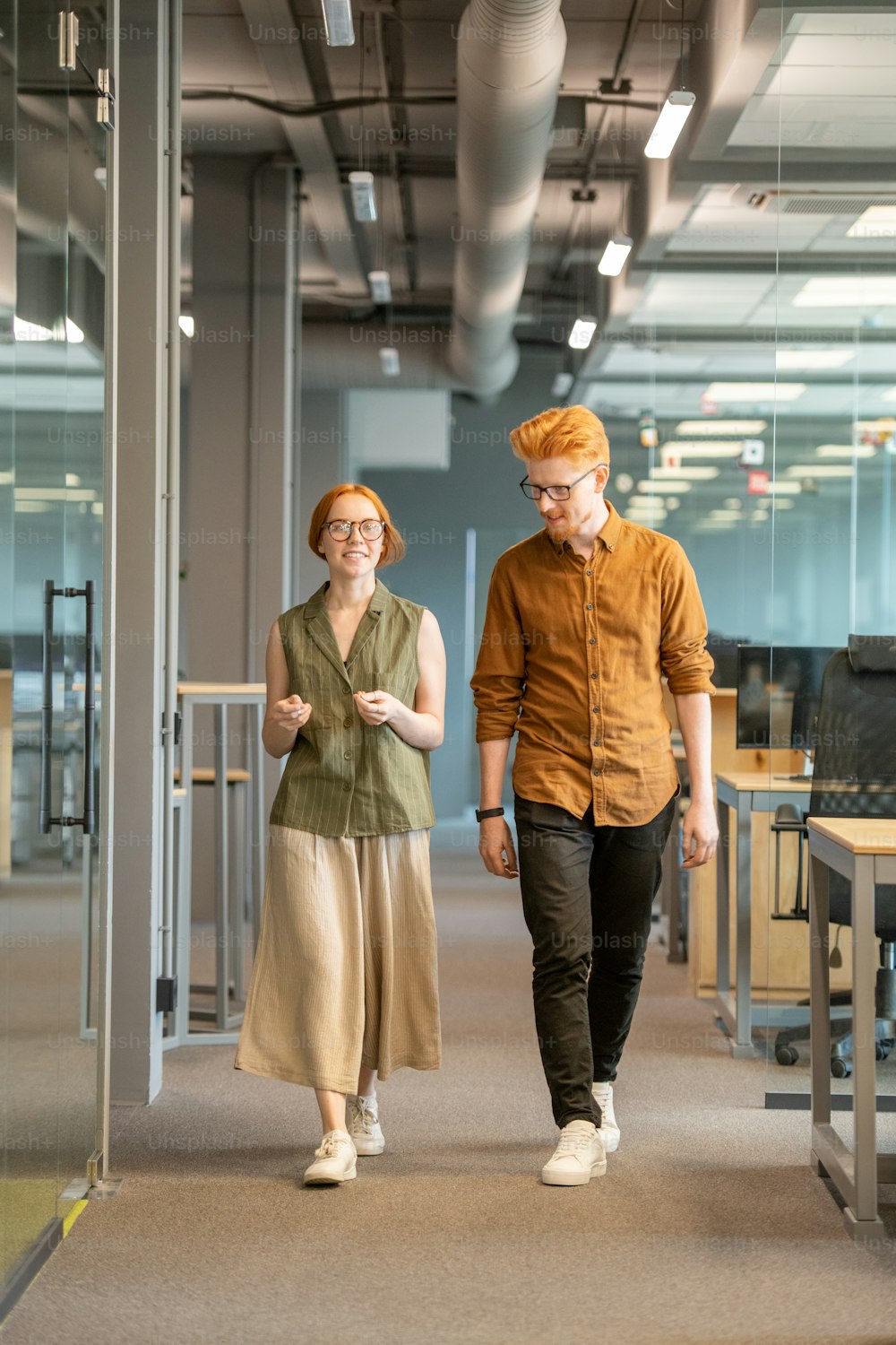 Happy young businesswoman in casualwear and her handsome colleague walking down aisle along row of desks and discussing work